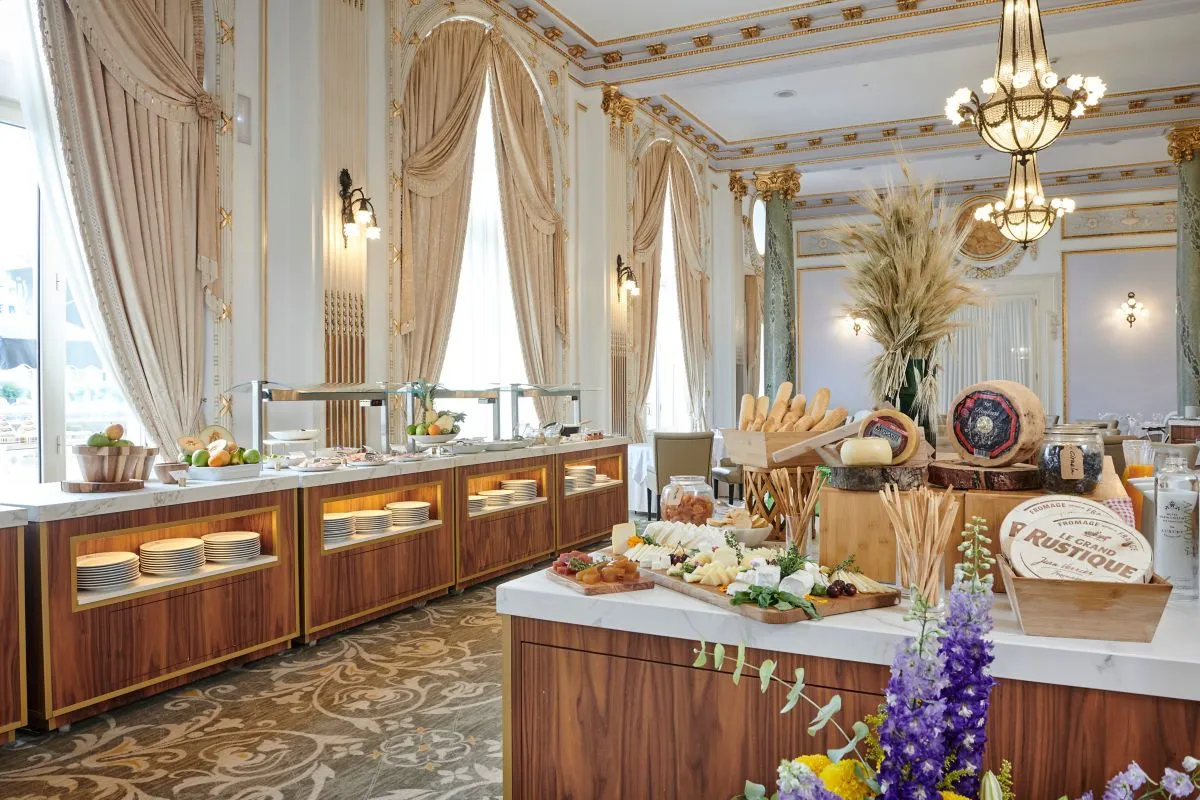 Otel Maria'nın modern ve konforlu bir yatak odasını sergileyen bu karede, beyaz nevresimlerle kaplı geniş bir yatak, şık bir yatak başlığı, iki yanında zarif komodinler ve pencereden süzülen doğal ışık, dinlendirici bir atmosfer sunuyor.