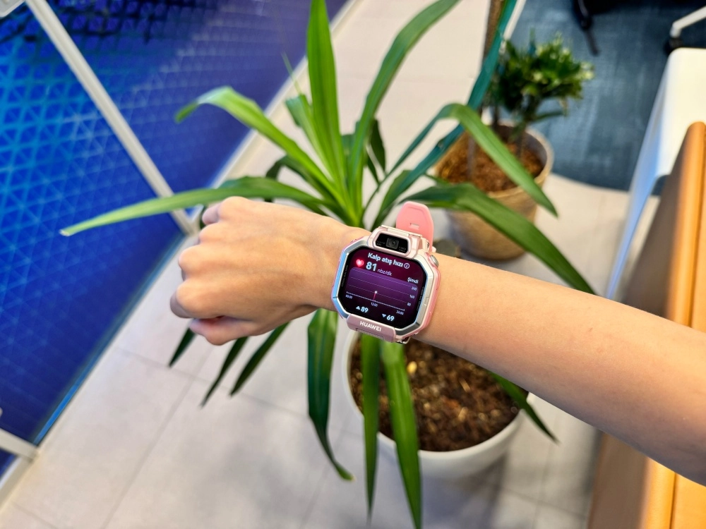 Bir kişinin kolunda, üzerinde Kalp atış hızı 81 nabız/dk gösterilen bir grafik ile nabız takibi verilerini sergileyen pembe renkli bir Huawei Watch Kids X1 akıllı saat görünüyor; arka planda saksı bitkileri ve mavi desenli bir duvar var.