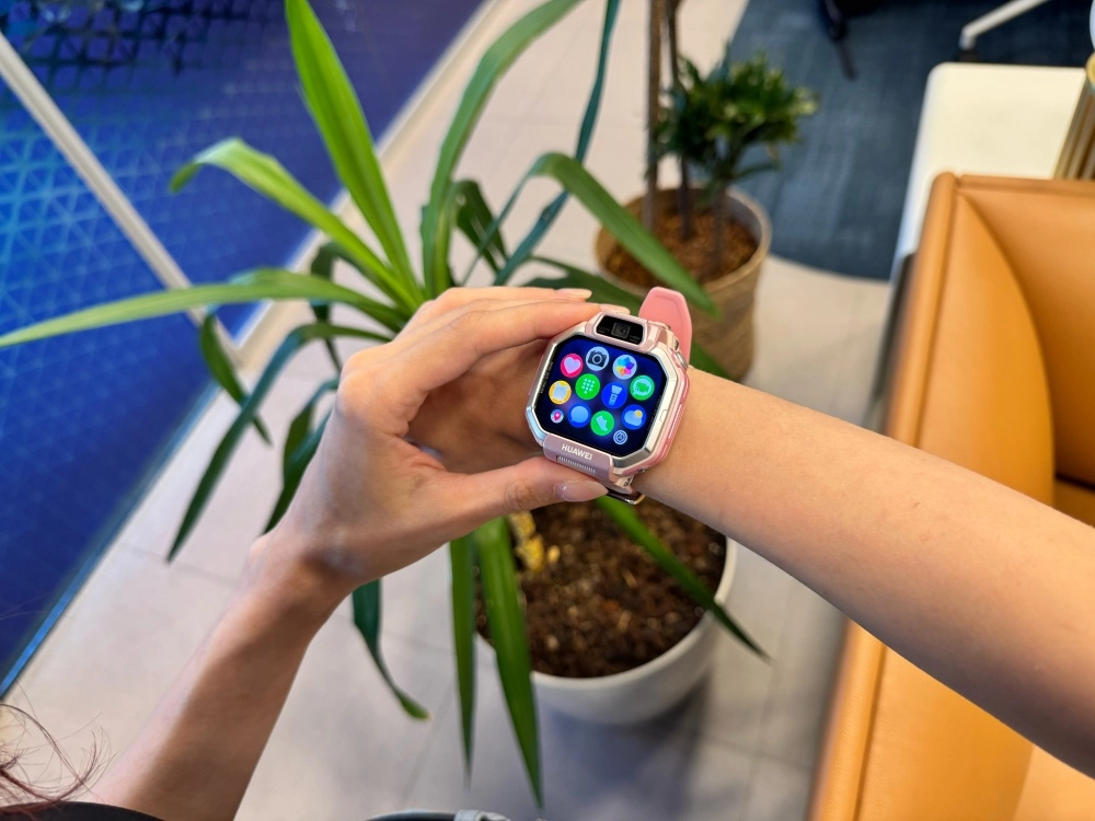 Genç bir kolun bileğinde duran pembe Huawei Watch Kids X1 akıllı çocuk saatinin, arkada bitkiler ve mobilyaların olduğu bulanık bir iç mekanda, renkli uygulama ekranı canlı bir şekilde görülüyor.