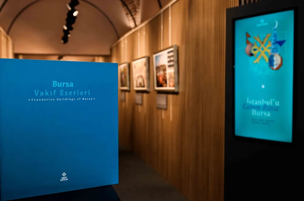Ön planda Bursa Vakıf Eserleri adını taşıyan büyük, mavi bir kitap, arkasında ahşap panelli bir duvar üzerinde sergilenen çerçeveli eserler ve sağda İstanbul'u Gören Rüya Bursa sergisini tanıtan dijital bir ekranın bulunduğu aydınlık bir galeri.