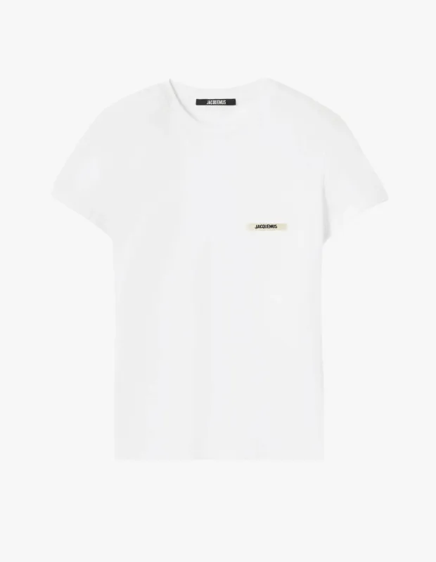 Açık gri bir zeminde düzgünce sergilenen, bisiklet yaka ve kısa kollu, minimalist tasarımlı Jacquemus beyaz tişörtün yaka kısmında siyah bir marka etiketi, göğsün sol tarafında ise krem rengi zemin üzerine yazılı Jacquemus logosu detayı net bir şekilde görülüyor.