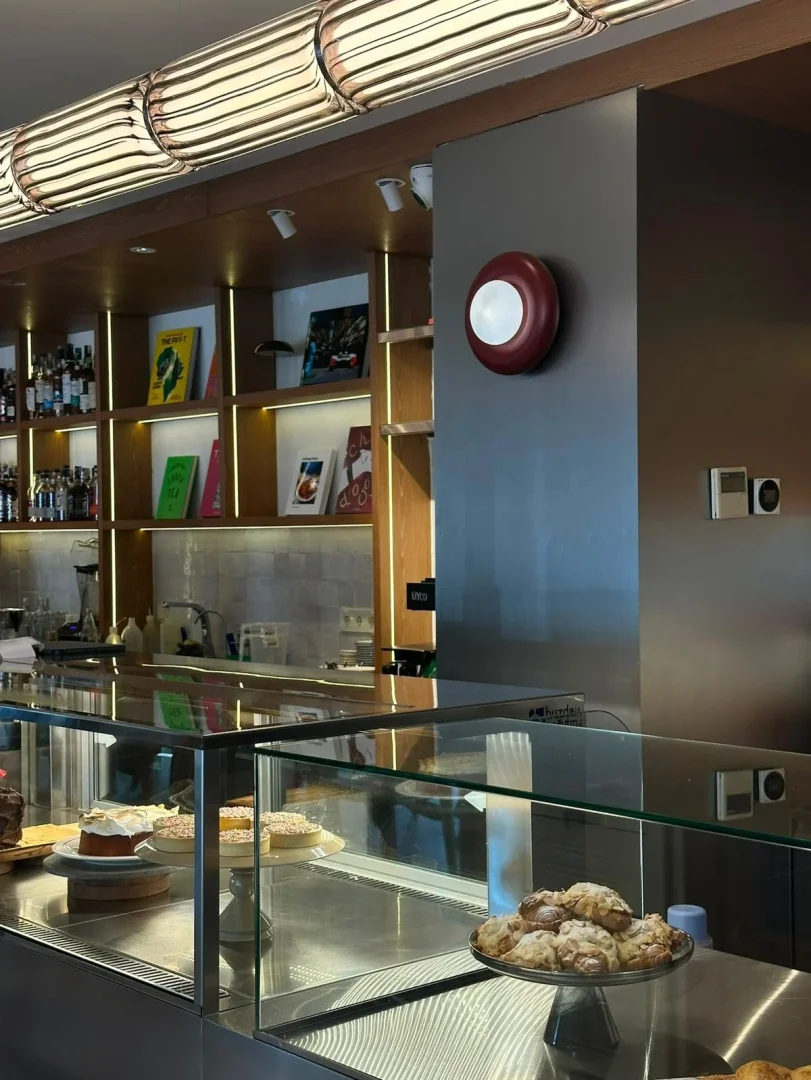Bu geniş iç mekanda, cam teşhir vitrininde göz alıcı pastaların ve tatlıların sergilendiği bir kafe veya bar görülürken, arkadaki aydınlatmalı ahşap raflar şişeler ve kitaplarla doludur; tavanın üst kısmındaki çizgili armatür ve duvardaki kırmızı yuvarlak ışık, modern tasarımı tamamlar.