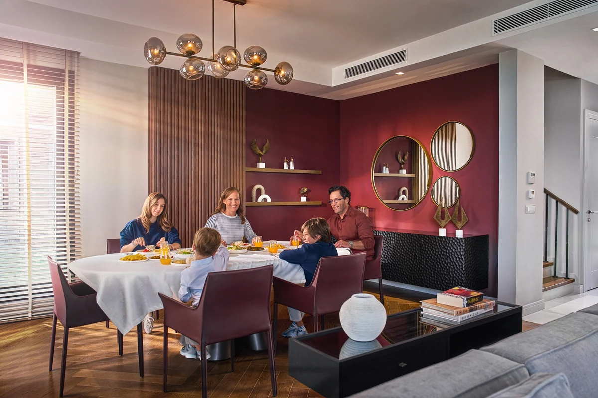 Modern bir kasaba evinin ferah ve aydınlık salonunda, farklı yaşlardaki aile üyeleri, özenle hazırlanmış yemeklerle dolu şık bir modern aile sofrasında keyifli bir akşam yemeği yiyor, samimi anlar paylaşıyorlar.