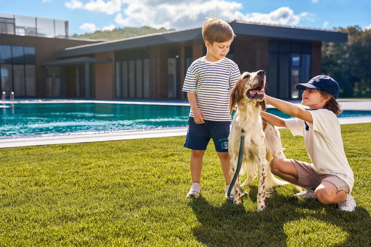 Kasaba Modern projesinin yemyeşil geniş çim bahçesinde, arkasında şık bir modern villa ve turkuaz havuz bulunan güneşli bir öğleden sonra, biri ayakta diğeri çömelmiş iki çocuk, tasmalı ve neşeyle bakan bir köpeği severek şehrin ortasında doğayla iç içe yavaş yaşamın huzurunu deneyimliyor.
