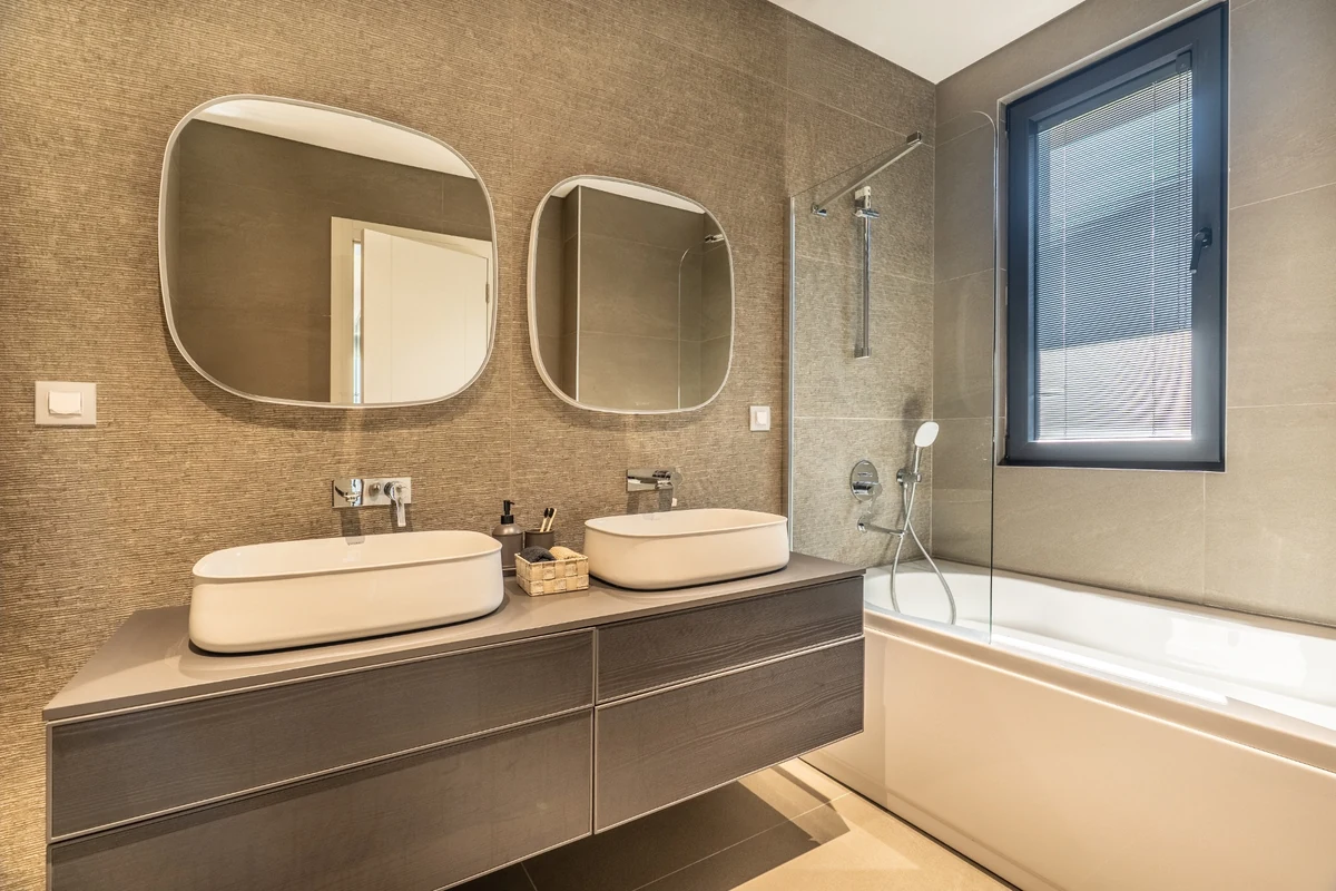 Geniş ve ferah bir his veren, açık renk tonlarının hakim olduğu modern bir üst kat banyo, büyük pencerelerden giren bol doğal ışıkla aydınlanırken, zarif ayaklı küveti ve minimalist ahşap detayları ile hem şık hem de işlevsel bir yaşam alanı olduğunu vurguluyor.