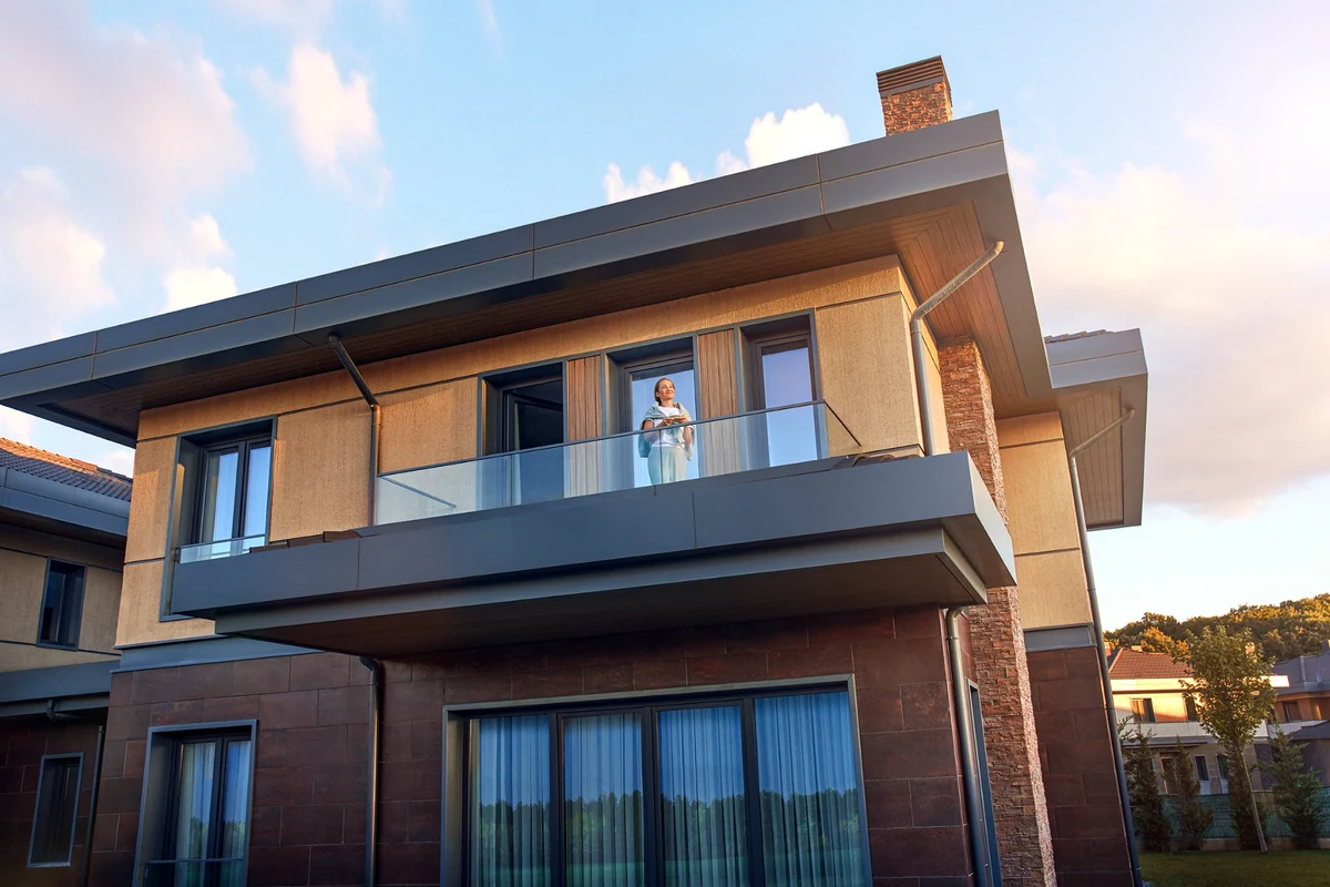 Bir Kasaba Modern villasının modern balkonunda günbatımı ışıkları altında elinde fincanıyla dışarıyı seyreden bir kadın, şehrin sunduğu imkânlardan vazgeçmeden yavaş yaşam felsefesini deneyimlemenin huzurunu yansıtıyor.