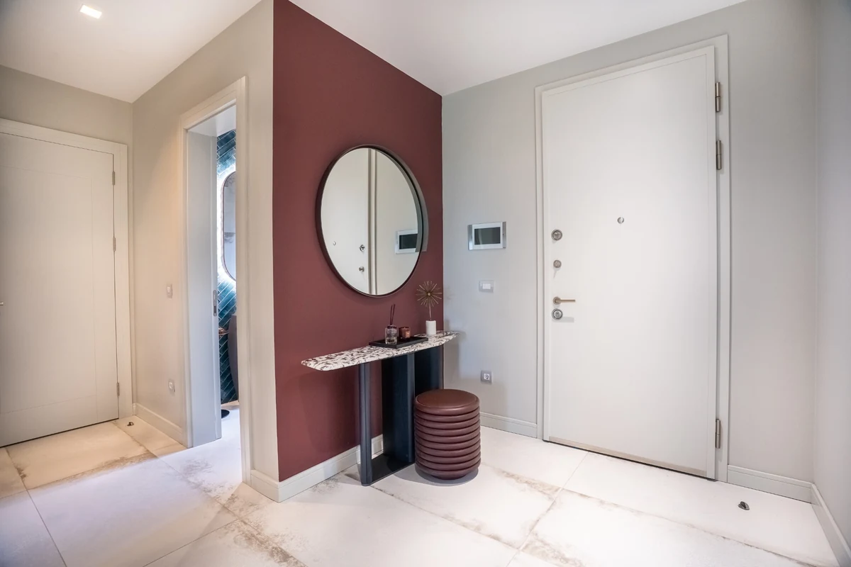 Beyaz ana kapılı ve açık gri duvarlı, zarif konsol ve tabure üzeri dairesel aynayla süslenmiş bordo renkli bir vurgu duvarının kontrast oluşturduğu modern bir villa giriş antresi, slow living yaklaşımı için vazgeçilmez bir karşılama alanı sunuyor.