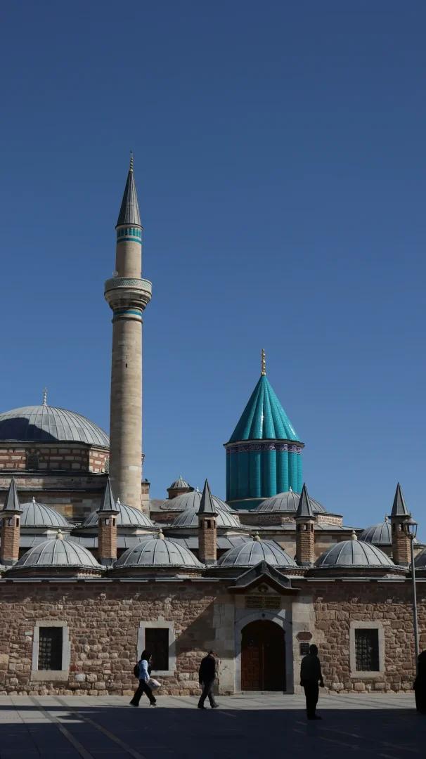 Güneşli bir günde, Konya'nın en önemli gezilecek yerlerinden Mevlana Müzesi'nin ikonik yeşil kubbesi, yüksek minaresi ve taş duvarlı yapıları önünde yürüyen ziyaretçiler, şehrin kültürel zenginliğini ve huzurlu atmosferini gözler önüne seriyor.