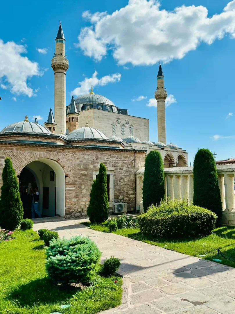 Konya'daki Mevlana Müzesi'nin yemyeşil bahçeleri arasından yükselen görkemli minareleri ve kubbeli yapılarıyla, berrak mavi gökyüzü altında huzurlu ve tarihi atmosferini gözler önüne seren geniş bir manzara.