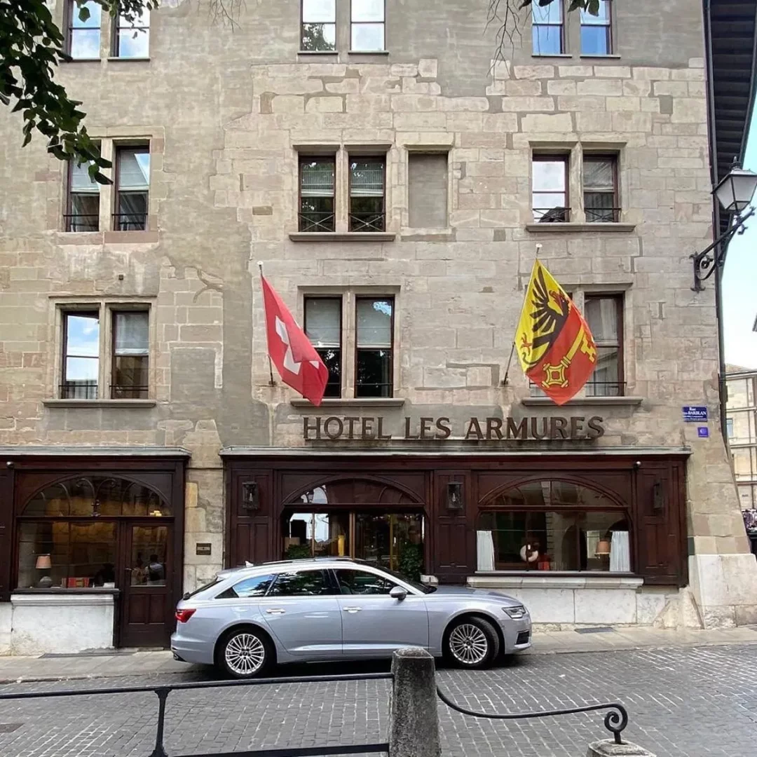 Cenevre'deki tarihi Hotel Les Armures'ın, İsviçre ve Geneva kantonu bayraklarıyla süslü, taş duvarlı ve koyu ahşap girişli cephesi önünde, gümüş renkli zarif bir station wagon Arnavut kaldırımlı bir sokakta park edilmiş halde, otelin klasik atmosferini modern bir dokunuşla tamamlıyor.