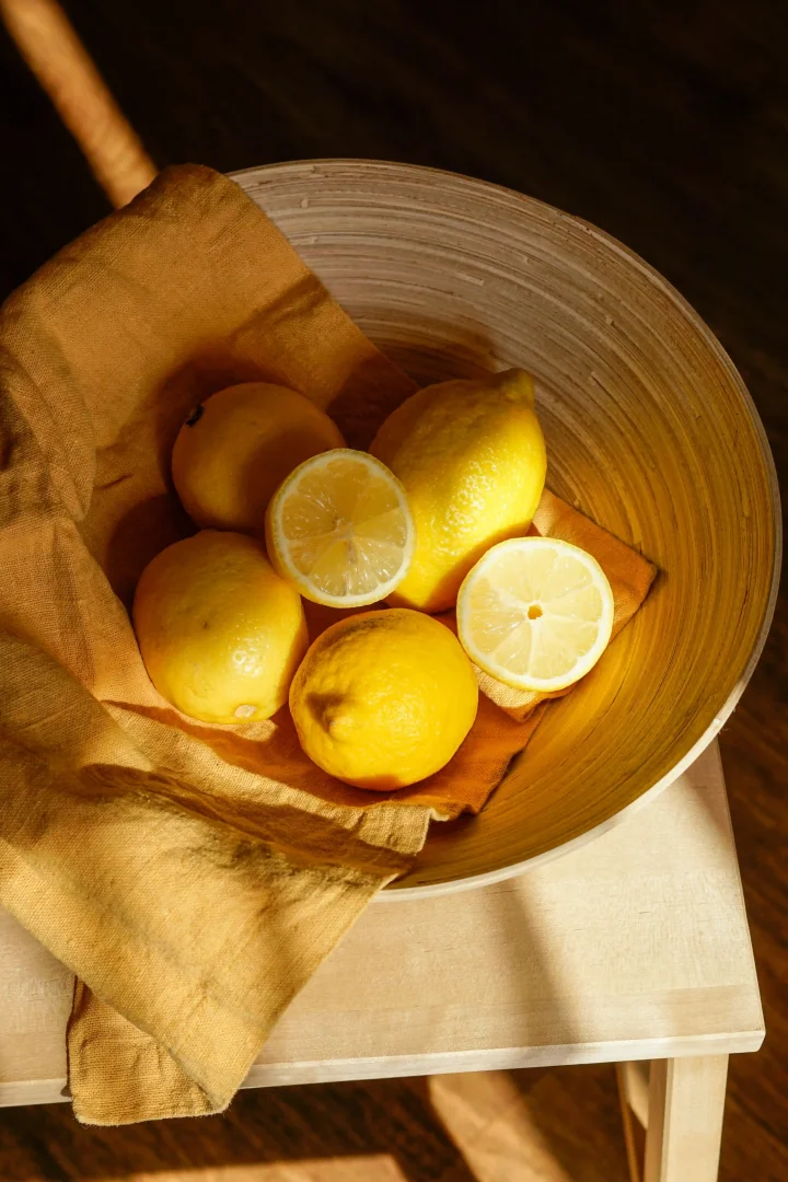 Ahşap desenli, derin bir kase içinde, güneş ışığıyla aydınlanmış, taze ve parlak sarı renkteki beş bütün limon ile iki yarım dilim limon, hardal sarısı keten bir bezin üzerinde sıcak bir şekilde duruyor.