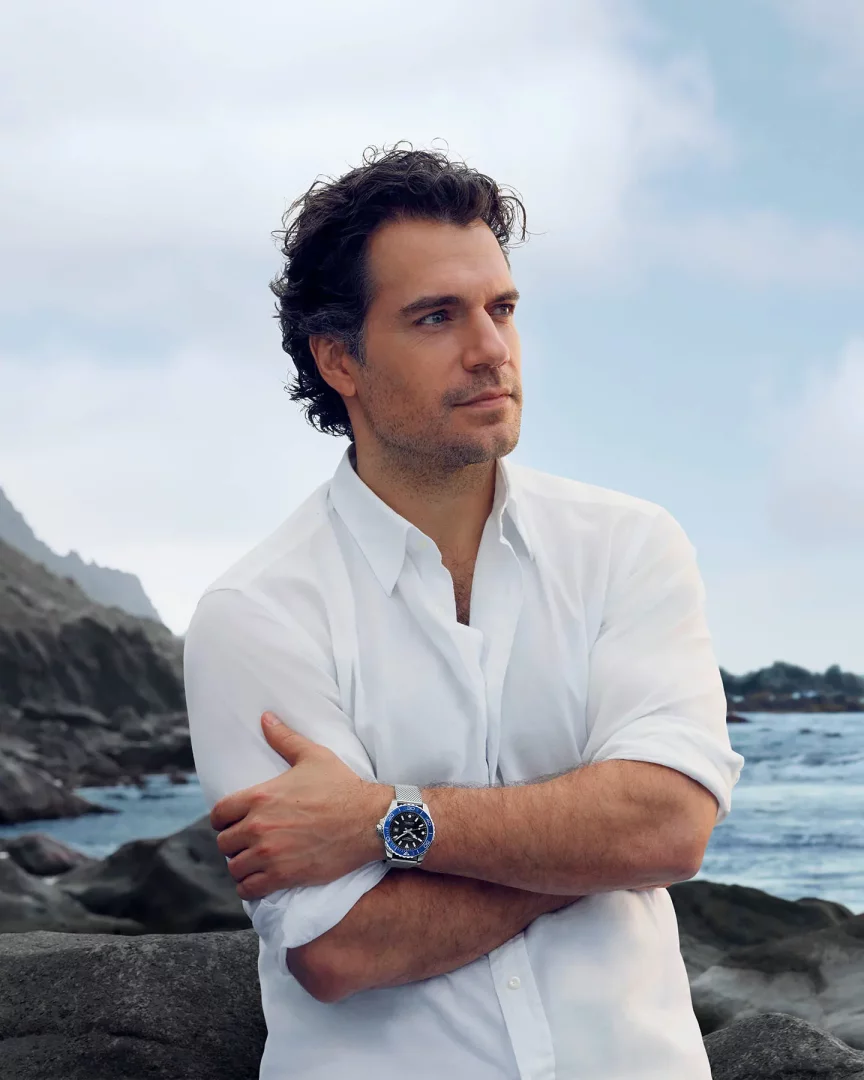 Henry Cavill, beyaz gömleği ve sol bileğindeki metal hasır kayışlı, mavi çerçeveli Longines saatiyle kollarını kavuşturmuş, kayalık bir sahil şeridinde dalgalı denize ve bulutlu gökyüzüne doğru bakıyor.