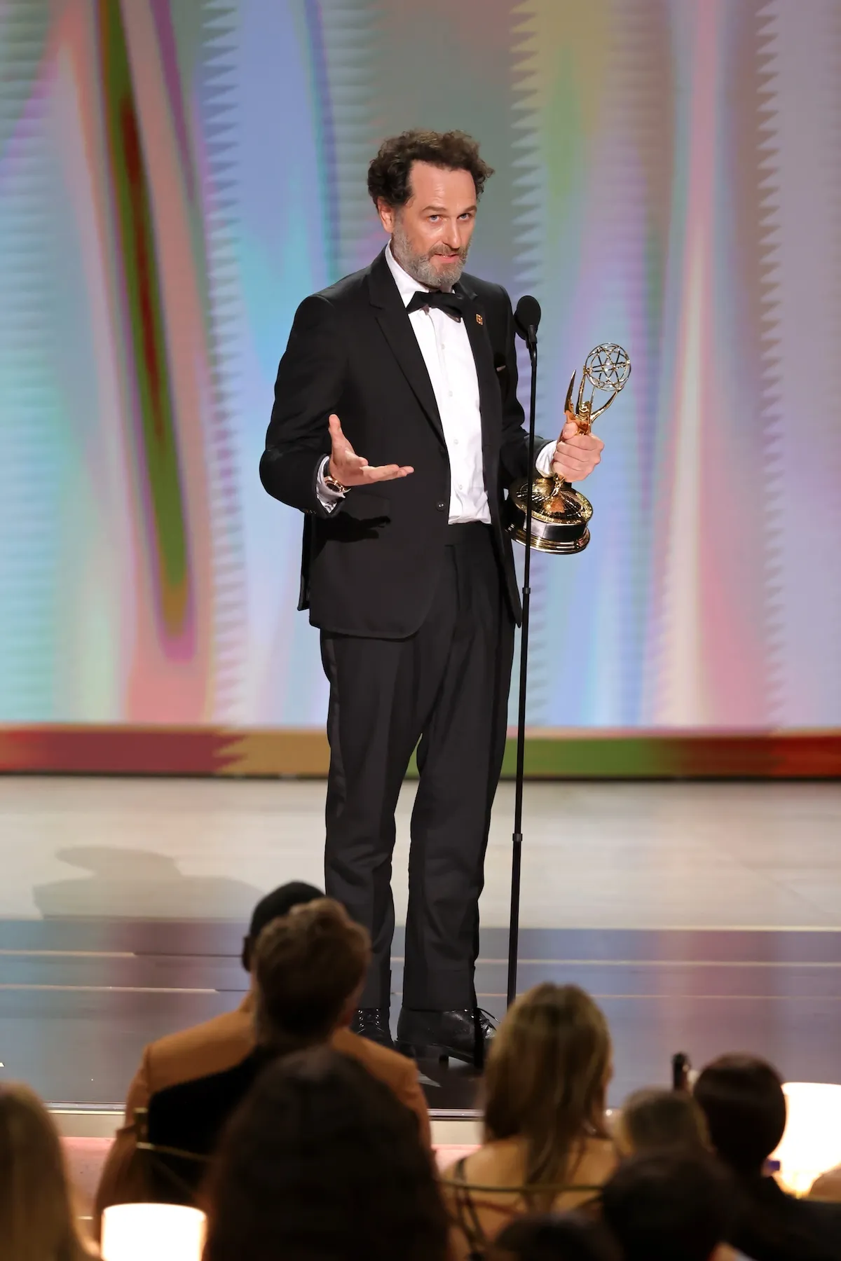 Matthew Rhys, siyah smokin ve papyonuyla sahnede, altın rengi bir Emmy ödülü tutarken sol eliyle jest yapıyor, arkasındaki renkli soyut arka plan ve önündeki seyirci kalabalığıyla çevrili bu anlamlı Emmy Ödülleri anını ölümsüzleştiriyor.