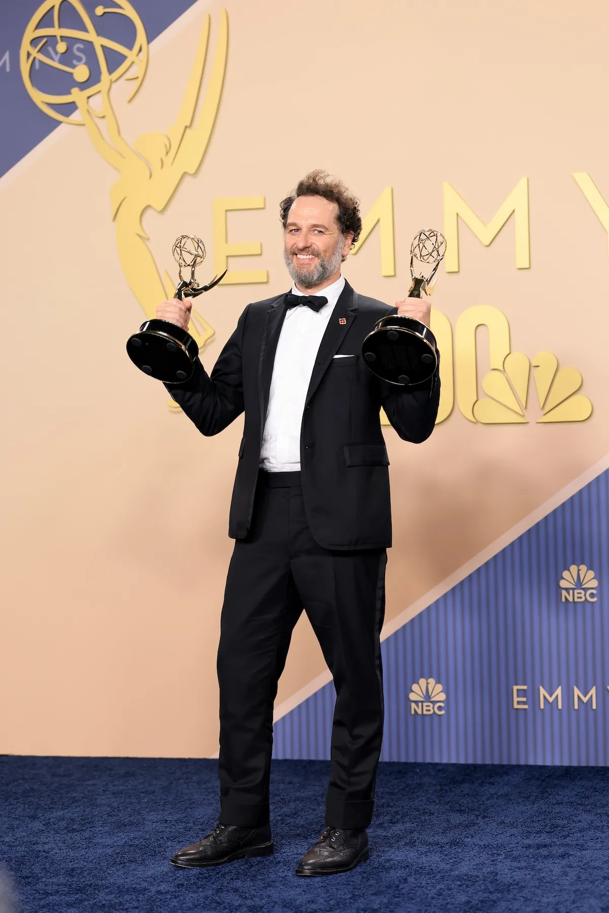 Matthew Rhys, mavi halıda dururken, şık siyah smokini ve papyonuyla gülümseyerek, her iki elinde de altın rengi iki Emmy ödülünü kaldırıyor ve arkasındaki büyük Emmy ve NBC logolarıyla uyumlu bir kompozisyon oluşturuyor.