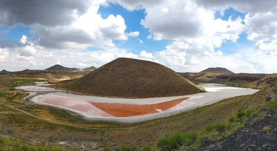Meke Krater Gölü'nün geniş açılı bir görüntüsünde, ortasında yükselen koyu kahverengi volkanik koni etrafında, sol kısımda pembe-kırmızı renkli sığ bir su birikintisi ve sağda daha açık gri renkte kısmen kurumuş bir göl yatağı belirginleşiyor; çevredeki seyrek otlu kahverengi tepeler ve gökyüzündeki parçalı bulutlar bu doğal oluşumun benzersiz silüetini ortaya koyuyor.
