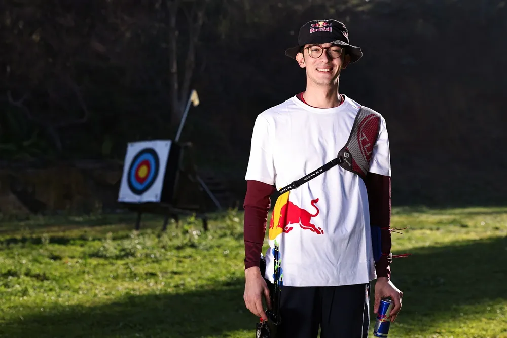 Gözlüklü ve Red Bull logolu şapka ile tişört giyen, üzerinde okçuluk göğüs koruyucusu bulunan ve elinde Red Bull kutusu tutan olimpiyat şampiyonu okçu Mete Gazoz, çim zeminde ok hedefinin önünde durarak gülümsemektedir.
