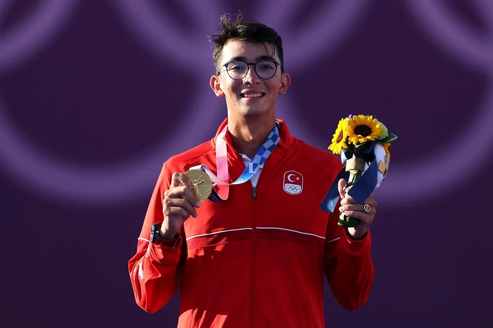 Mete Gazoz, yuvarlak gözlükleri ve kırmızı milli takım formasıyla, boynundaki altın madalyayı sağ eliyle tutarken gülümseyerek kameraya bakıyor, sol elinde tuttuğu ayçiçekleriyle kariyerinin ve hayatının en parlak anlarından birini gururla sergiliyor.