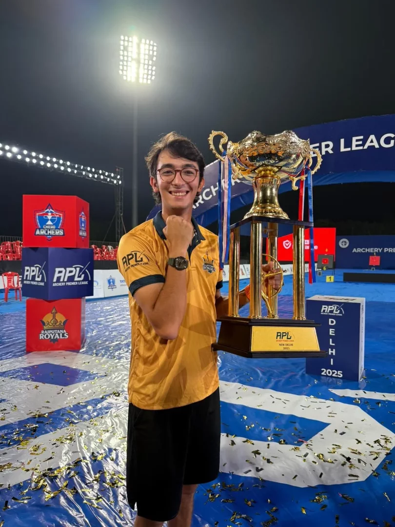 Olimpiyat şampiyonu milli okçumuz Mete Gazoz, Archery Premier League afişleri ve APL New Delhi 2025 yazılarının görüldüğü, mavi zemin üzerinde altın konfetilerin saçıldığı bir spor alanında, büyük altın bir kupayı kaldırarak kameralara gülümseyen mutlu bir poz veriyor.