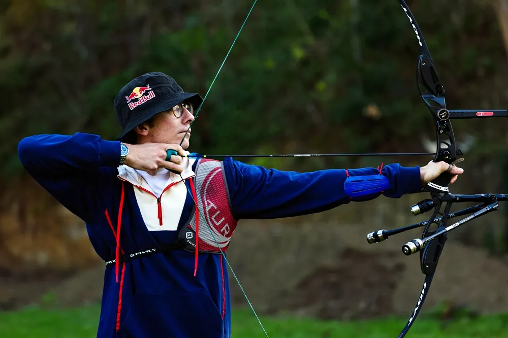 Red Bull logolu siyah şapkası, gözlükleri ve mavi sweatshirt'üyle Türk okçuluk sporcusu Mete Gazoz, bir ok atışı sırasında recurve yayını gergin bir şekilde tutarak hedefine kararlılıkla odaklanmış, arka planda yeşil ağaçlıklar seçiliyor.