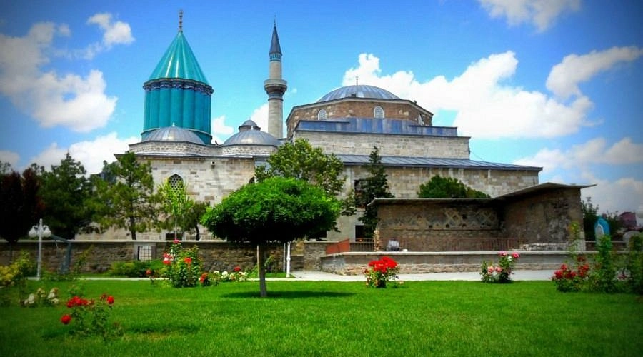 Konya'daki Mevlana Müzesi'nin ikonik yeşil kubbesi ve minaresi, ön planda yemyeşil çimenler, çeşitli ağaçlar ve kırmızı güllerle bezeli huzurlu bir bahçe içinde, berrak mavi gökyüzü altında tüm ihtişamıyla yükseliyor.