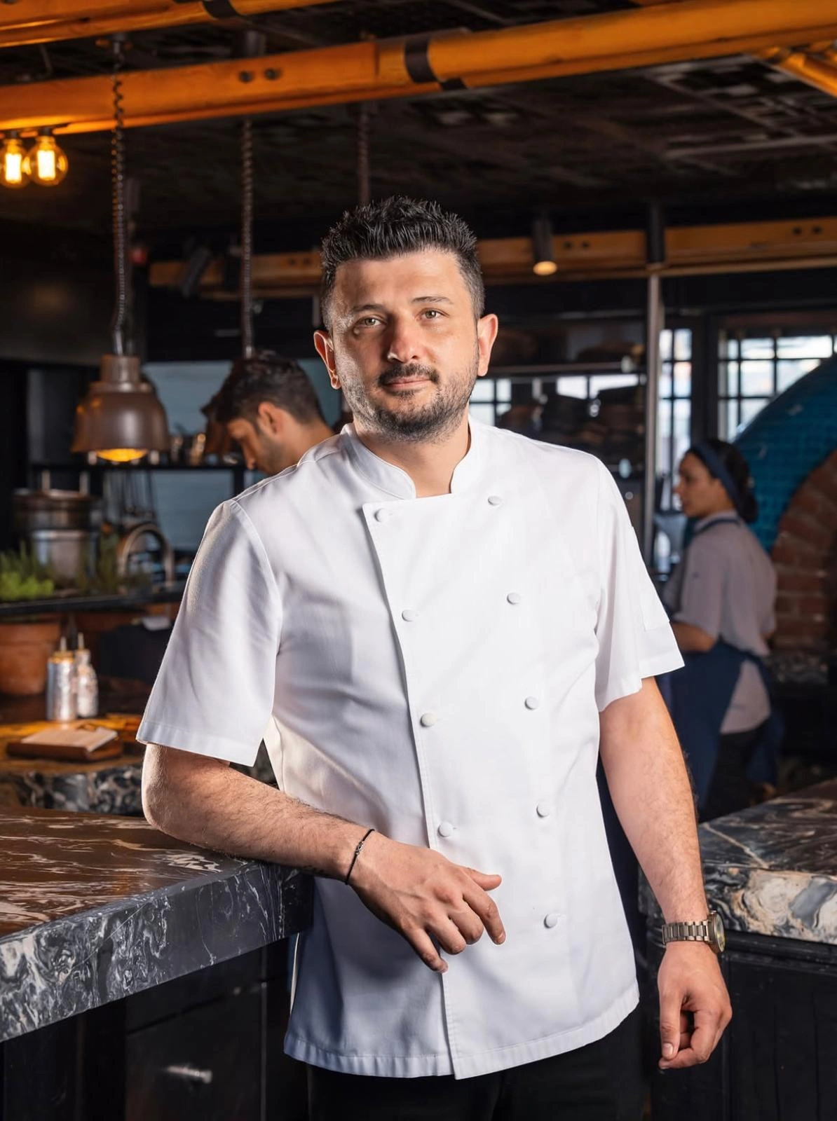 Şef Mevlüt Özkaya, beyaz aşçı ceketini giymiş, kalabalık bir restoran mutfağında koyu renkli mermer bir tezgaha yaslanarak kameraya doğrudan bakıyor; arkasında çalışanlar ve endüstriyel aydınlatmalar modern ve hareketli bir ortamı vurguluyor.