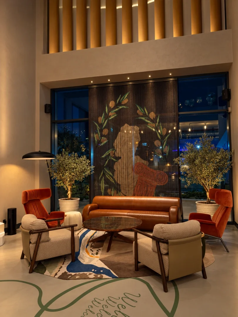 Otel lobisinde yer alan bu modern ve konforlu dinlenme alanında, şık bir deri kanepe, çeşitli renklerde yumuşak koltuklar, zeytin dalları motifli büyük bir dekoratif perde ve ortada ahşap bir sehpa bulunurken, zemindeki Hoşgeldiniz yazısı ve saksı bitkileri mekana sıcak bir atmosfer katıyor.
