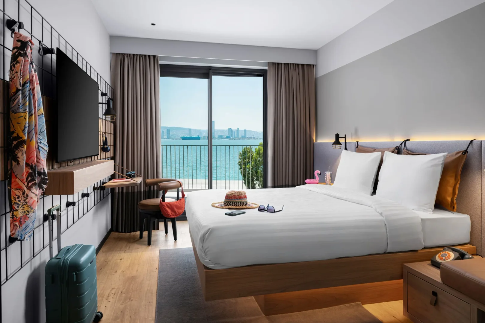 Modern bir Moxy otel odası, büyük bir deniz manzarası sunan cam kapıya bakan, üzerinde hasır şapka ve güneş gözlüğü bulunan beyaz çarşaflı konforlu bir yatakla dolu; sol duvarda ise renkli bir şal ve televizyon ünitesiyle birlikte şık bir depolama alanı dikkat çekiyor.