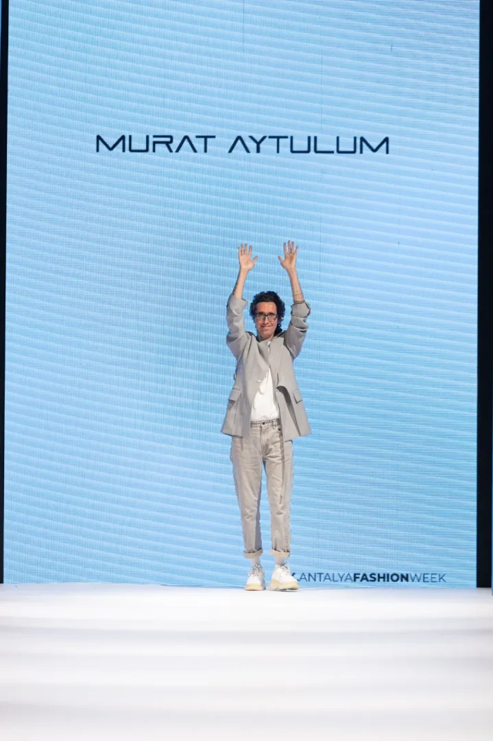Tasarımcı Murat Aytulum, Antalya Moda Haftası podyumunda, arkasında kendi adının yazılı olduğu devasa mavi bir ekranın önünde, krem rengi takım elbisesi ve spor ayakkabılarıyla elleri havada izleyicileri selamlayarak duruyor ve etkinliğin atmosferini yansıtıyor.