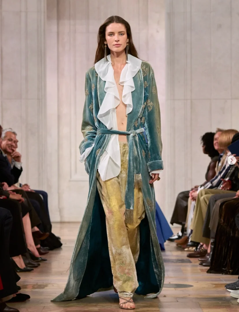 Ralph Lauren'in New York Moda Haftası defilesinde, işlemeli teal kadife bir robdöşambr, beyaz fırfırlı bir bluz ve desenli altın rengi pantolon giyen bir model podyumda zarif adımlarla yürürken, seyirciler dikkatle izliyor.