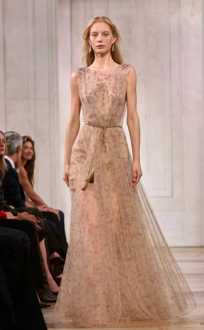 Ralph Lauren defilesinde, New York Moda Haftası podyumunda yürüyen sarışın bir model, belinde püsküllü ince bir kemer ve zarif soluk pembe çiçek desenleriyle süslenmiş, yere kadar uzanan, şeffaf tül bir elbiseyle büyüleyici bir duruş sergiliyor.