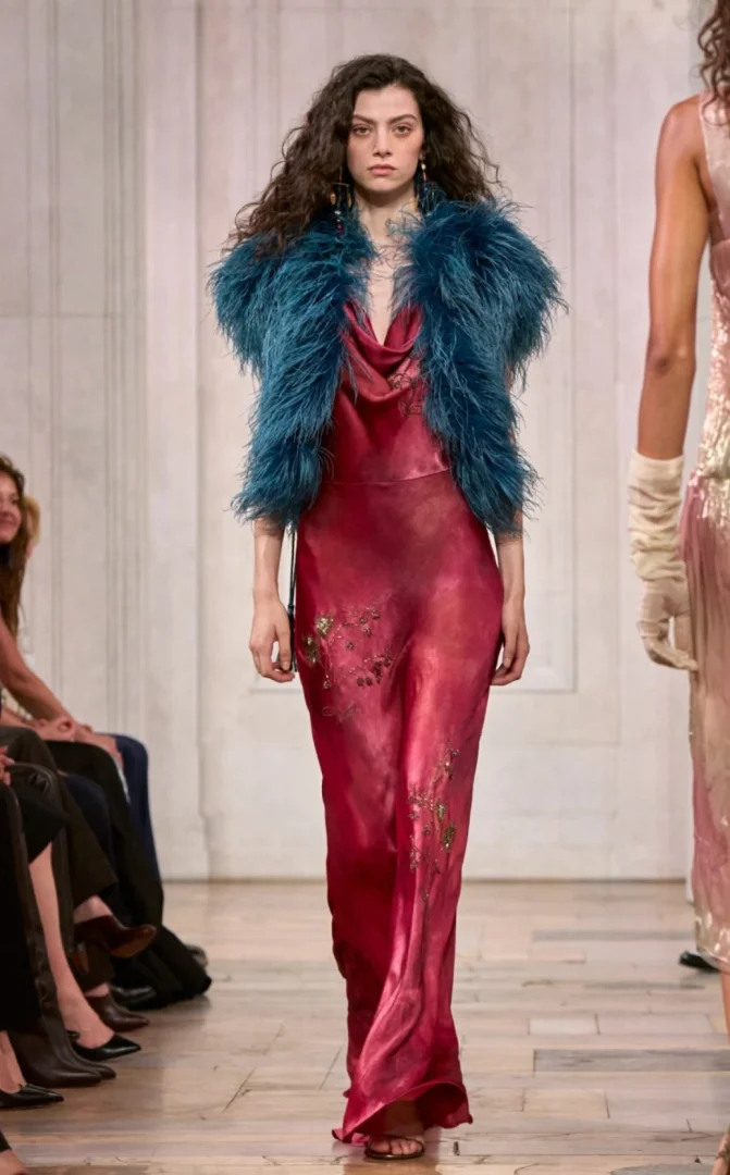 New York Moda Haftası kapsamındaki Ralph Lauren defilesinde, altın işlemelerle süslenmiş parlak fuşya rengi askılı bir elbise ve canlı turkuaz tüylü kısa bir ceket giyen, uzun dalgalı koyu saçlı bir model podyumda yürüyor.
