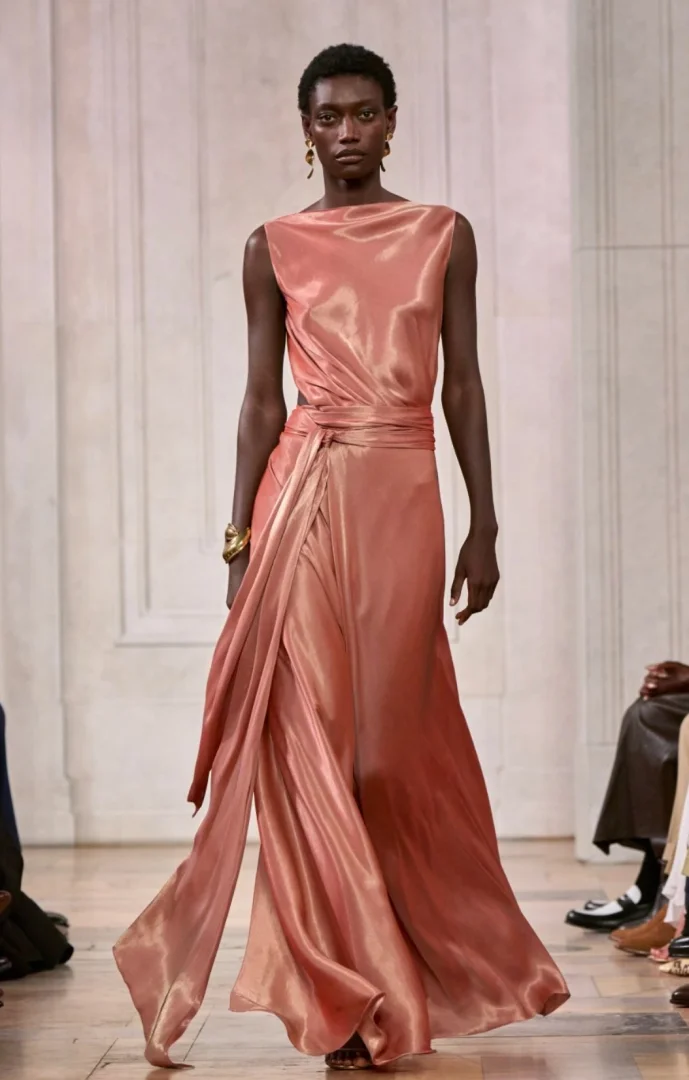 New York Moda Haftası kapsamında Ralph Lauren defilesinde podyumda yürüyen model, şeftali pembe tonlarında parıltılı, uzun, drapeli ve belden kuşaklı ipek bir elbise giyiyor, altın küpeler ve bilekliklerle şıklığını tamamlıyor.