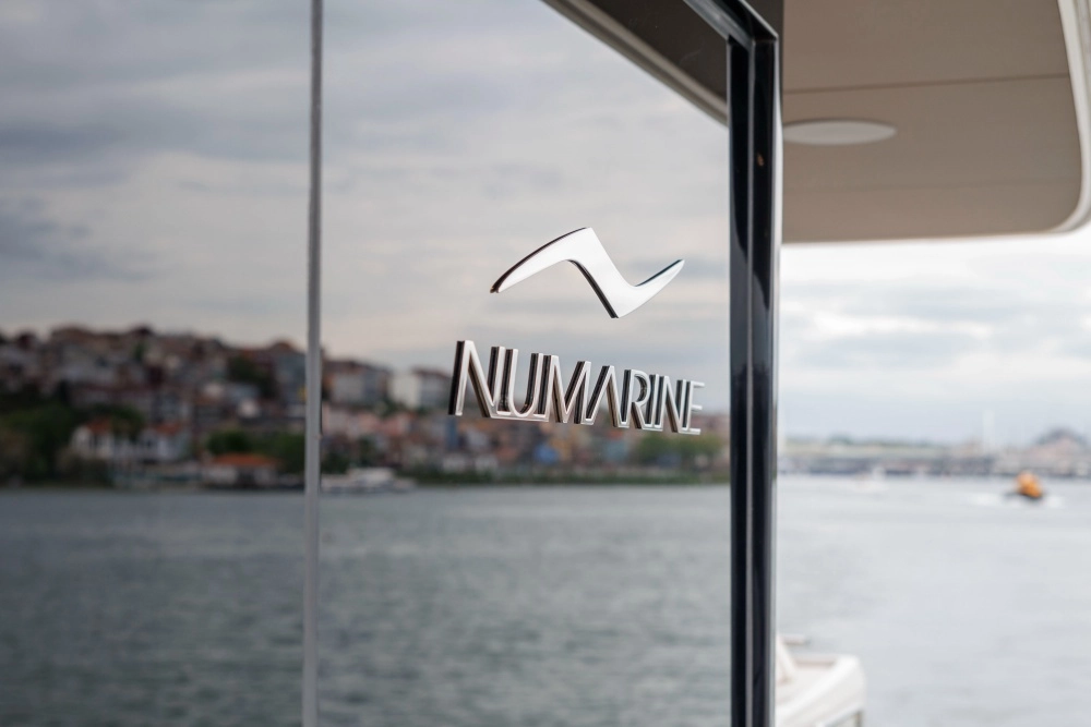 Gümüş renkli Numarine logosu ve markası, yansıtıcı bir yat penceresinde belirgin bir şekilde yer alırken, bulanık arka planda kıyı şeridi evleri ve dingin deniz manzarası Numarine'in denizcilikteki uzmanlığını ve lüks yat deneyimini vurguluyor.