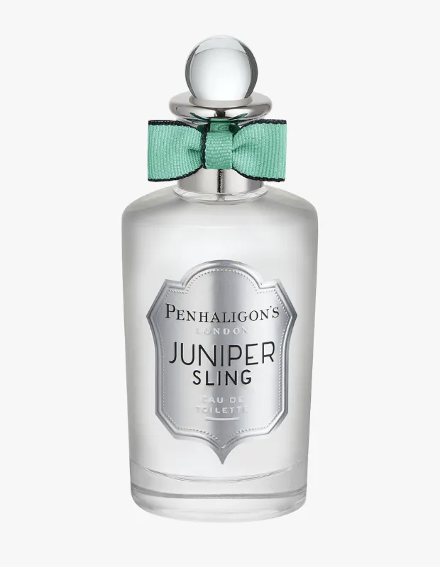 Penhaligon's Juniper Sling parfüm şişesi, koyu renkli etiketinde beyaz yazılar, gümüş yuvarlak kapağı ve boynundaki açık renkli kurdele ile dikkat çekerken, içindeki sarımsı kehribar sıvı şeffaf camından görünen bulanık, açık renkli bir arka plan önünde zarif bir şekilde konumlanmıştır.