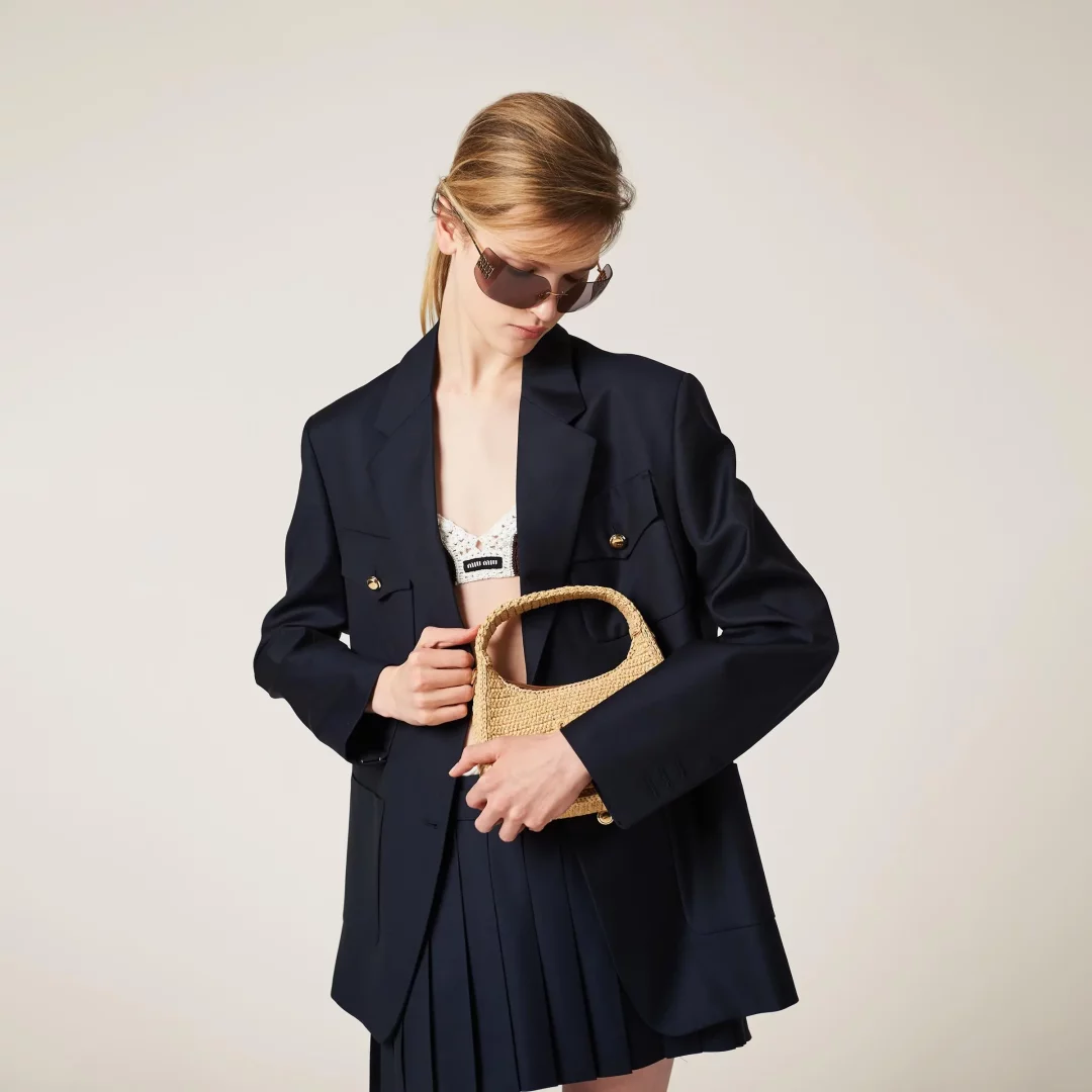 Sarı saçları arkadan toplanmış, koyu renk güneş gözlüklü bir model, altın düğmeli lacivert bol bir blazer ceket, Miu Miu logolu dantel sütyen ve pileli etekle modern bir preppy stilini yansıtırken, zarif hasır çantasını dikkatlice tutuyor.
