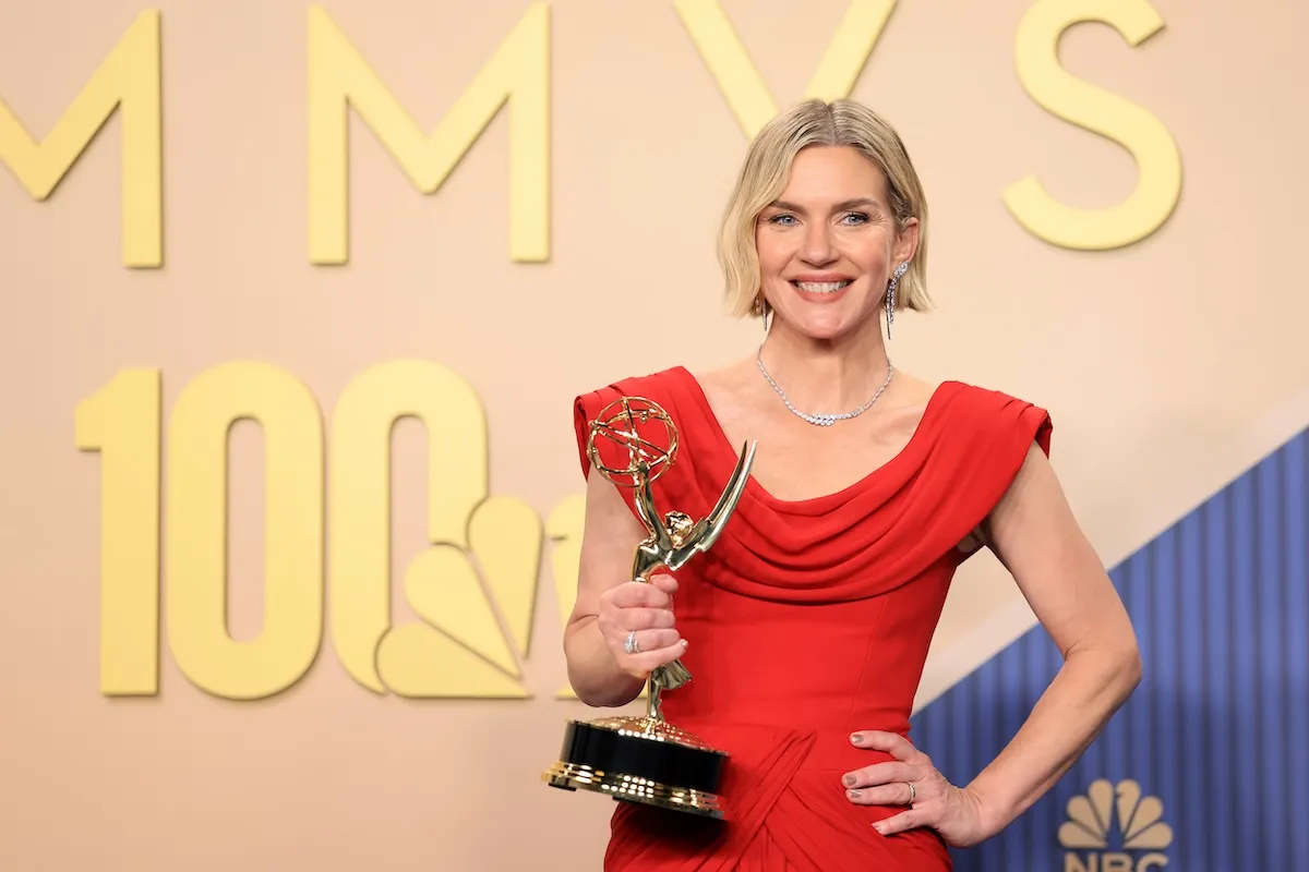 Rhea Seehorn, zarif kırmızı, omuzları drapeli bir elbiseyle, elmas küpeleri ve kolyesiyle gülümsüyor, sol elinde parıldayan bir Emmy ödülü tutarak poz veriyor; arka planda, Emmy Ödülleri törenini çağrıştıran altın rengi büyük harfler ve NBC logosu belirgin.