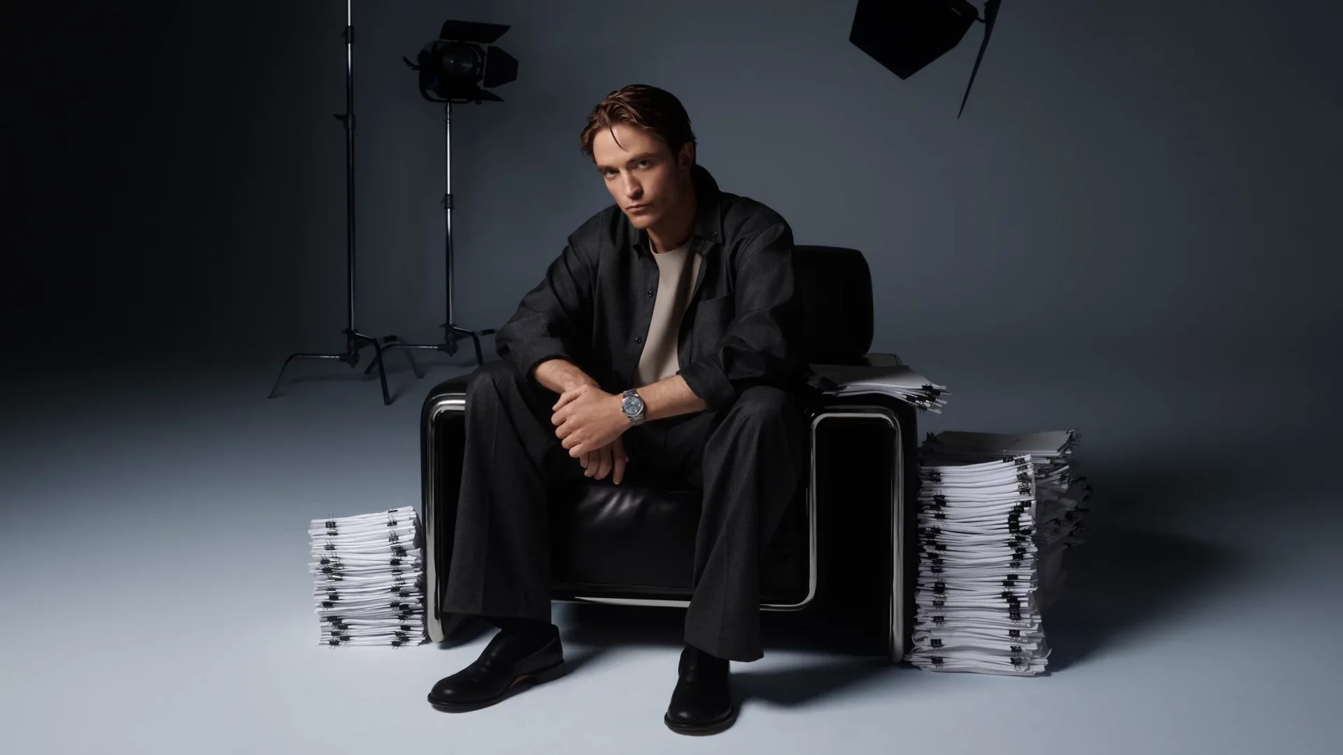 Robert Pattinson, stüdyo fonunda siyah deri bir koltukta, iki yanında dikkat çekici yüksek kağıt desteleriyle oturmuş, koyu renk giysileri ve sol bileğindeki şık metal saatiyle ciddi bir ifadeyle doğrudan kameraya bakıyor.