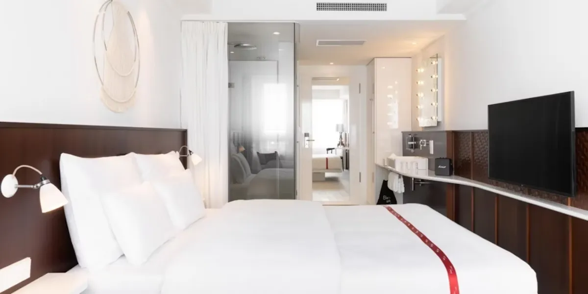Beyaz nevresimli ve koyu renk ahşap başlıklı yatağın hâkim olduğu bu modern Ruby Cenevre otel odasında, yatağın solunda duvara monte aydınlatmalar ve dekoratif bir duvar süsü yer alıyor; sağ tarafında ise ahşap panelli bölümde lavabo, aynalı makyaj ışıkları, siyah Marshall hoparlör ve büyük bir televizyon modern bir bütünlük sunarken, arka planda cam bölmeli banyo alanı ve başka bir odaya açılan kapı odanın ferah düzenini gösteriyor.