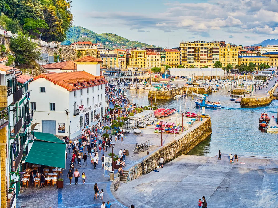 San Sebastian şehrinin ikonik La Concha Körfezi'nin parlayan turkuaz sularını, uzun kumsalı boyunca uzanan hareketli şehir binalarını ve güneşin aydınlattığı geniş plajında dinlenen insanları gösteren, şehrin enerjisini yansıtan büyüleyici bir genel görünüm.