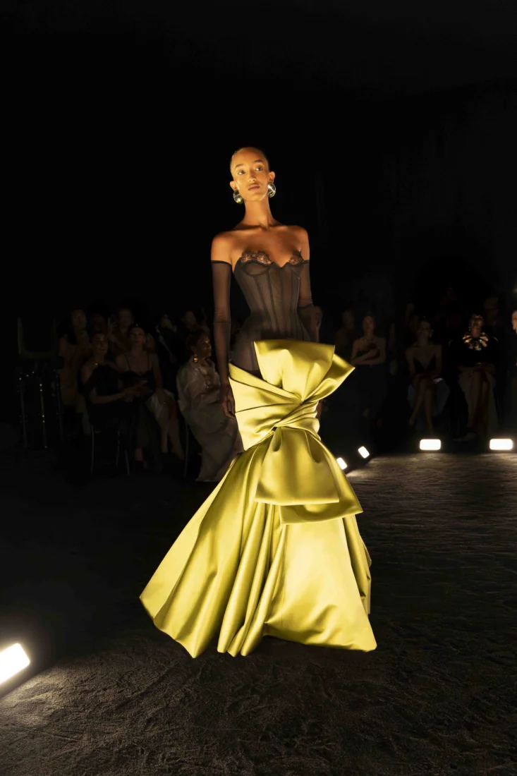 Model, Schiaparelli haute couture koleksiyonundan, siyah dar bir kıyafetin üzerine giyilmiş, karın kaslarını vurgulayan, altın rengi, heykelsi bir göğüs zırhı ile podyumda güçlü bir duruş sergiliyor.