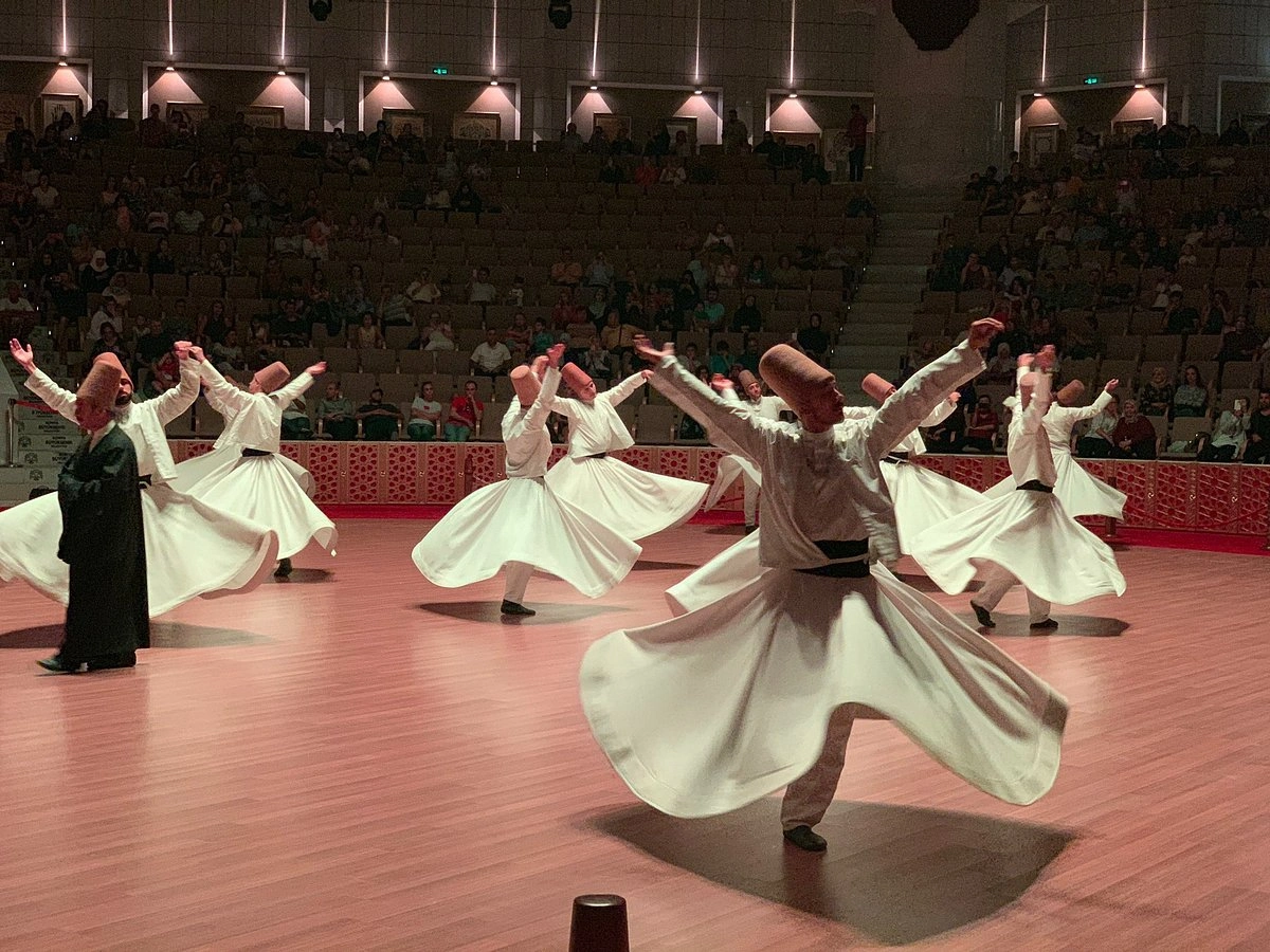 Geniş bir salonda parlatılmış ahşap zeminde, beyaz tennureleri ve kahverengi sikkeleriyle birkaç semazen, kolları açık ve etekleri savrularak Sema töreni gerçekleştirirken, onları çevreleyen tribünlerdeki kalabalık izleyici topluluğu bu mistik performansı hayranlıkla izliyor.