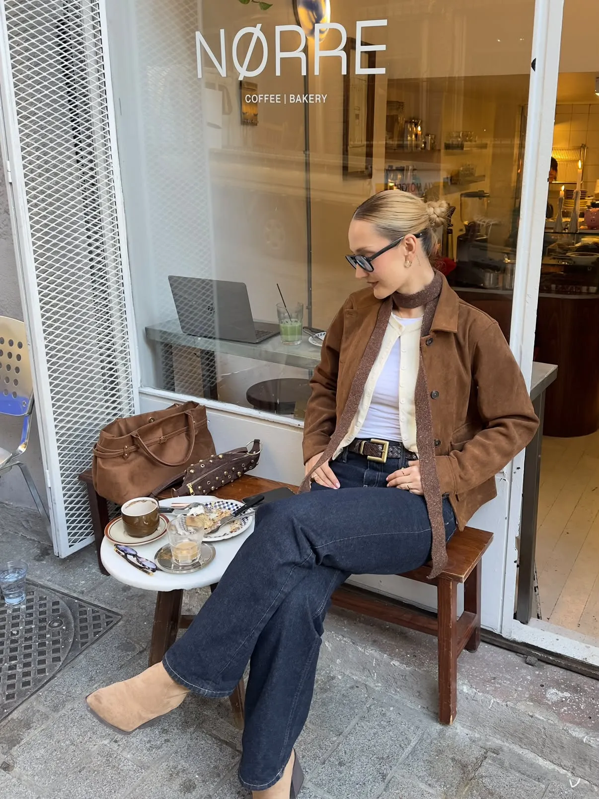 NØRRE COFFEE | BAKERY tabelasının altında, şehir hayatının keyfini çıkaran stil sahibi bir kadın, kahverengi süet ceketi, koyu kot pantolonu ve güneş gözlükleriyle dışarıda oturmuş, küçük masasında duran kahvesi ve tatlısıyla keyifli bir kahve molası veriyor.