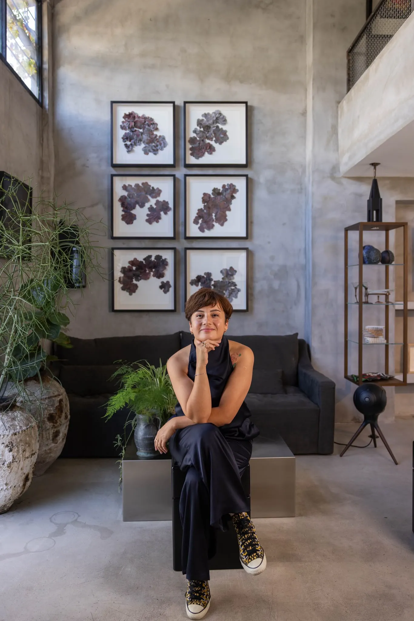 Sevim Kaya, modern sanat eserlerinin sergilendiği bir iç mekanda, doğal formlardaki altı çerçeveli tablo ile gri beton duvar önünde koyu renkli kıyafetleri ve desenli spor ayakkabılarıyla metal bir küp üzerinde oturmuş, kameraya gülümserken.