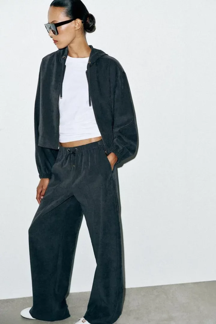 Beyaz duvarda koyu gri kapüşonlu üstü, beyaz crop top'u, siyah güneş gözlüğü ve bol paça rahat pantolonuyla poz veren kadın, seyahat için konforlu ve şık bir görünüm sergileyen bu pantolonun pratikliğini vurguluyor.