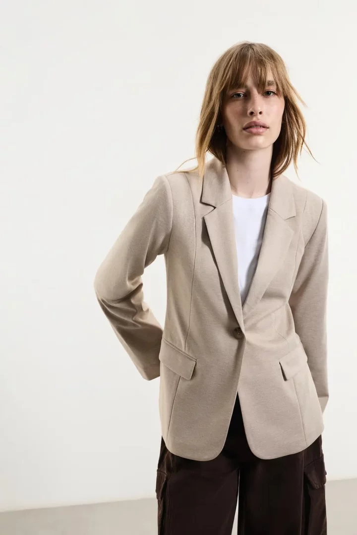 Genç bir kadın, beyaz tişört ve koyu renk kargo pantolon üzerine giydiği açık bej rengi blazer ceketiyle beyaz bir arka plan önünde duruyor, seyahat stil rehberi için pratik ve şık bir blazer kombini sunarak zarafet ve rahatlığı birleştiriyor.