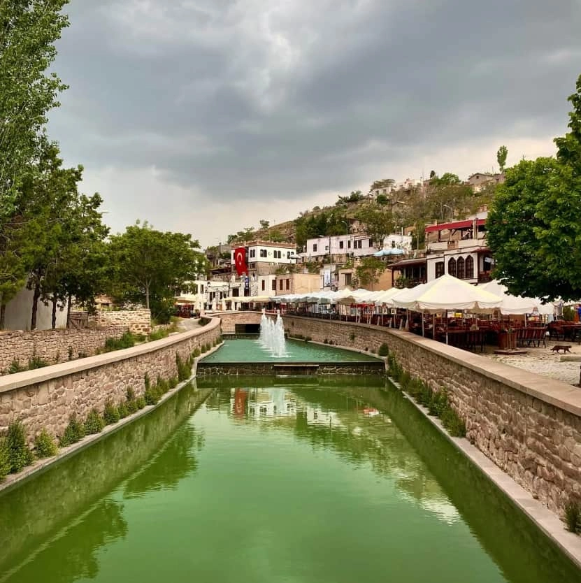 Sille Köyü'nde, gri ve bulutlu bir havada, ortasında yemyeşil suyun aktığı ve fıskiyelerin yükseldiği taş duvarlı bir kanalın, sağ tarafında beyaz şemsiyelerle donatılmış açık hava kafelerinin, sol tarafında yeşil ağaçların ve tepenin yamacına yayılan Türk bayraklı geleneksel beyaz evlerin göründüğü genel bir manzara.
