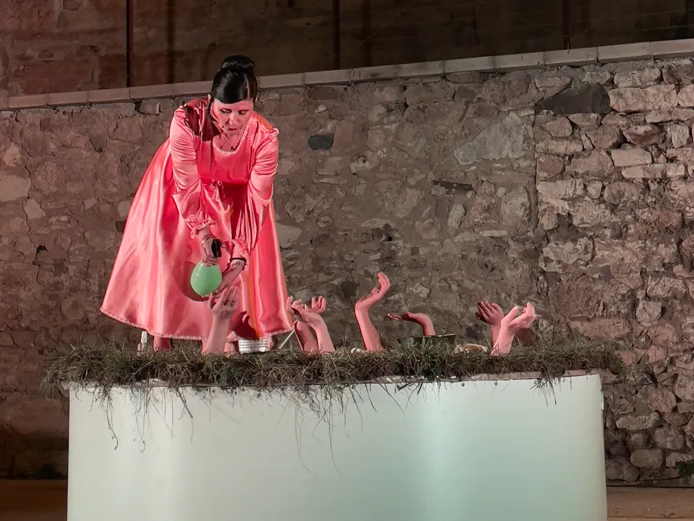 Pembe, parlak elbiseli bir kadının Sinop Bienali'nde sergilenen bir performans sanatında, samanla kaplı beyaz silindir bir yapıdan uzanan birçok cansız elin üzerine yeşil bir sprey şişesinden dikkatlice bir sıvı püskürttüğü, taş duvarlı bir mekanda çekilmiş etkileyici bir görüntü.