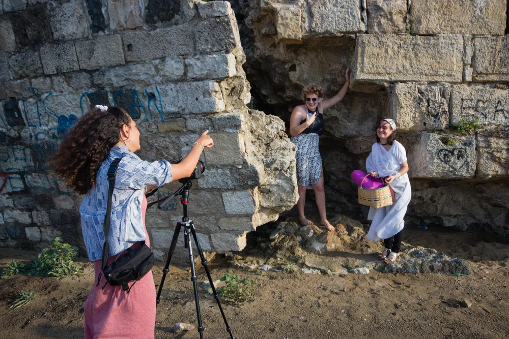 Sinop Bienali kapsamında, tarihi taş duvarların ve kumlu toprağın önünde gerçekleşen bir sanatsal performansta veya fotoğraf çekiminde, kıvırcık saçlı bir yönetmen kamerası başında iki farklı tarzdaki kadın performansçıyı yönlendirirken, biri modern kıyafetleri içinde duvara yaslanmış, diğeri ise beyaz tuniğiyle elinde hasır sepet tutarak gülüyor.