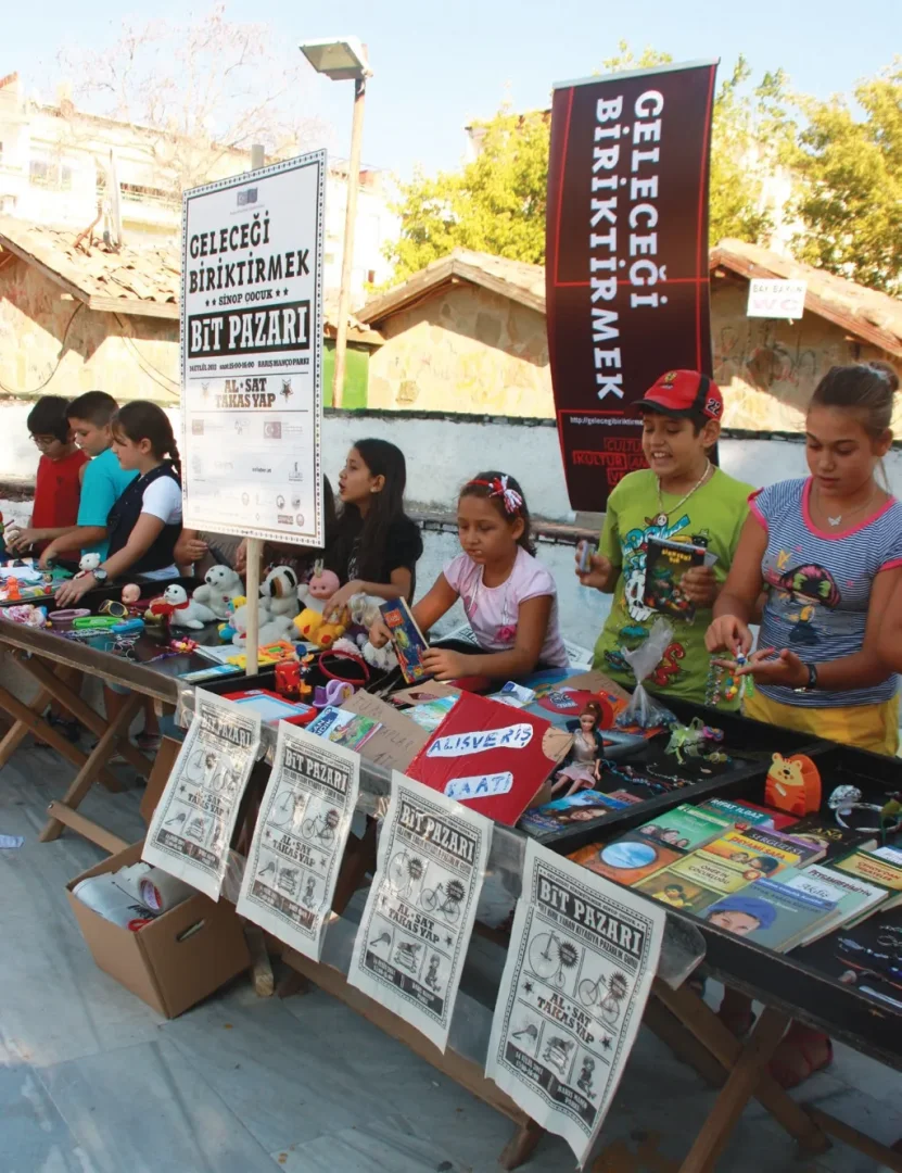 Sinop Bienali kapsamında düzenlenen, Geleceği Biriktirmek temalı bir açık hava Bit Pazarı'nda, farklı yaşlardaki çocuklar tezgahların başında ikinci el oyuncaklar, kitaplar ve çeşitli eşyaları alıp satarak veya takas ederek toplumsal katılım ve sürdürülebilir tüketim pratiği sergiliyorlar.