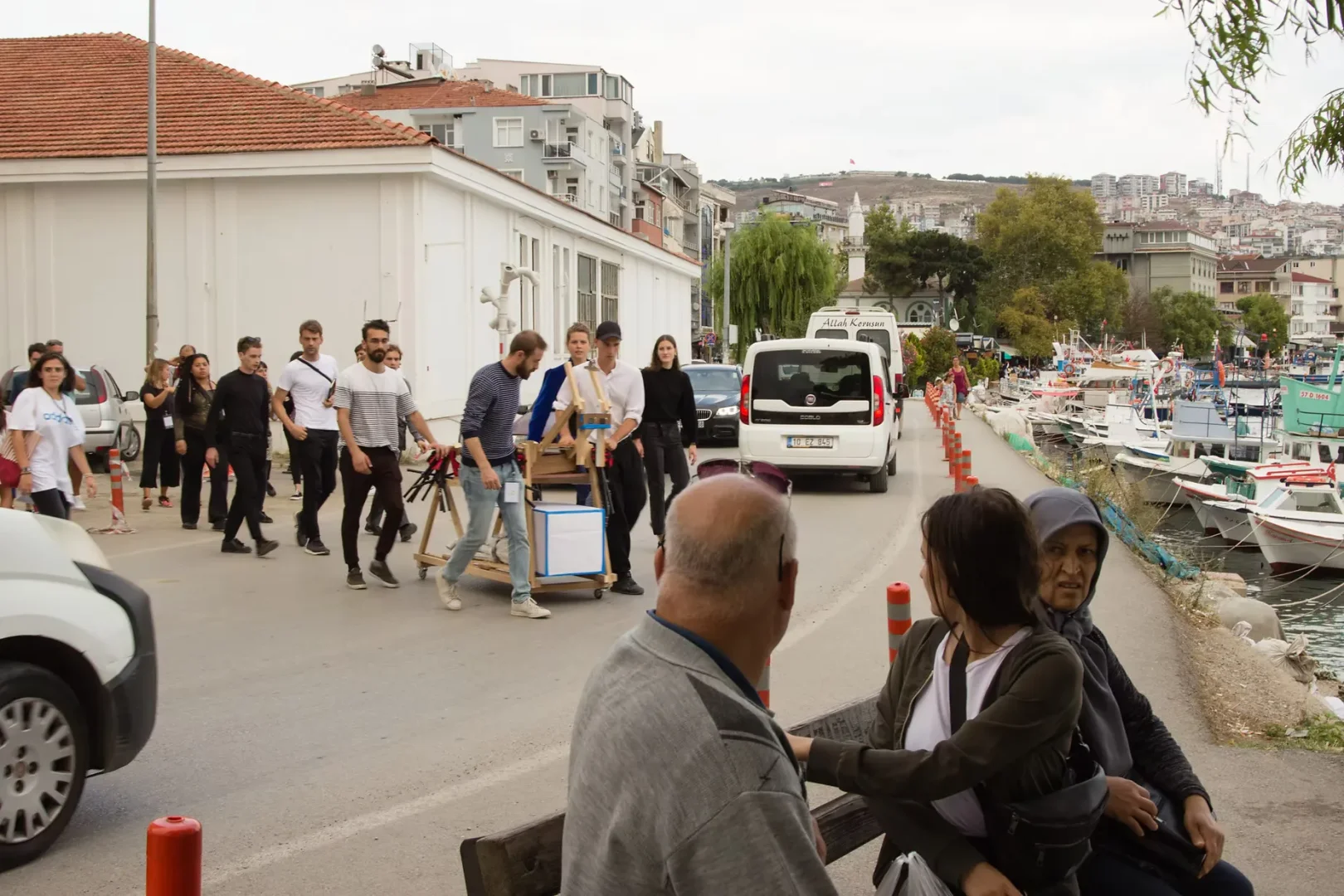 Sinop Bienali etkinlikleri kapsamında, Sinop'un hareketli liman çevresinde, ahşap bir el arabası ve çeşitli ekipmanlarla yürüyen gençlerden oluşan bir grup, beyaz binaların ve tepedeki yerleşim yerlerinin fon oluşturduğu kalabalık bir caddede ilerlerken, rıhtıma dizilmiş tekneler ve ön planda bankta oturanlar dikkat çekiyor.